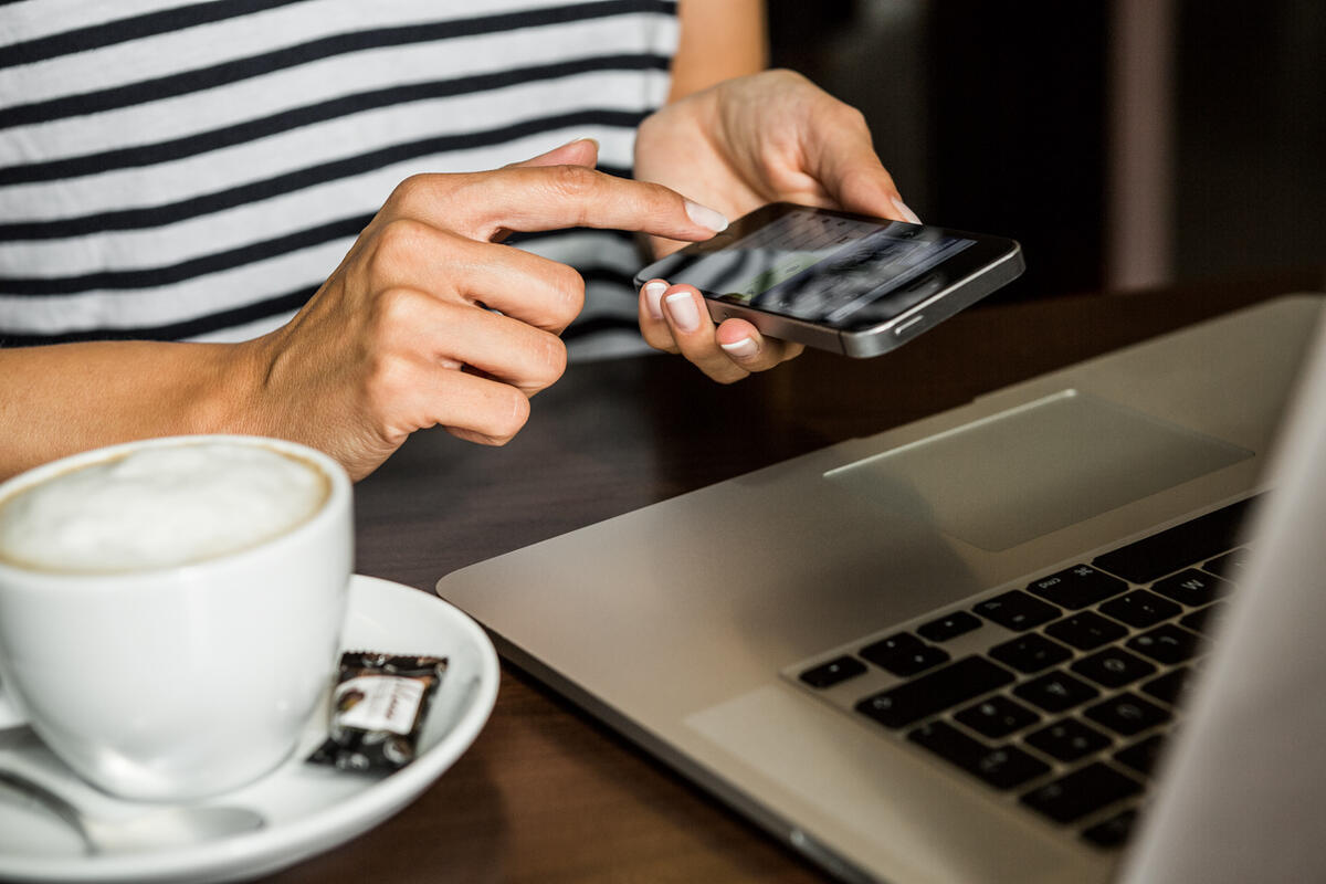 Person bedient Smartphone an Schreibtisch mit Laptop und Kaffeetasse